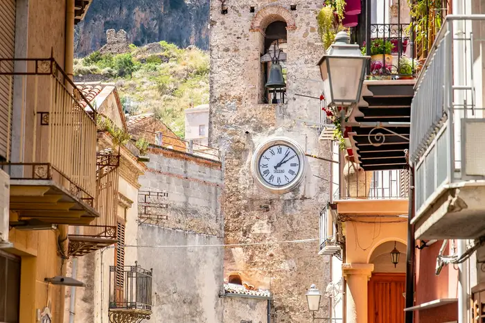 A narrow Sicilian street with a stone bell and clock tower below a rocky hillside